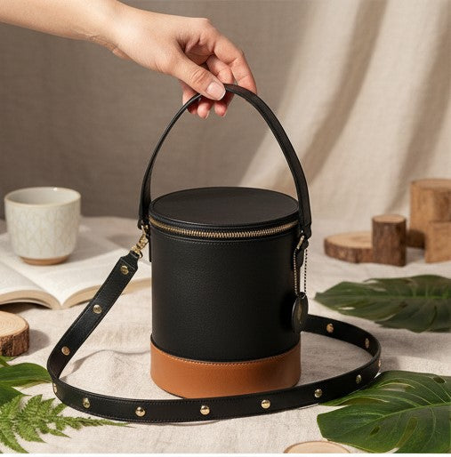 Hand holding the Hemera plant leather bucket bag in black and gingerbread, featuring a structured cylindrical silhouette, top handle, detachable shoulder strap, and contrast colour base.