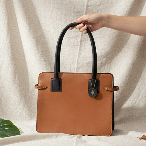 Aranyani plant-based leather tote bag in gingerbread and black colour held by hand, structured silhouette with dual handles against a neutral fabric background