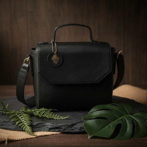 Asteria black plant-based leather crossbody bag with structured silhouette placed on a textured surface with green leaves, showcasing a premium sustainable handbag design
