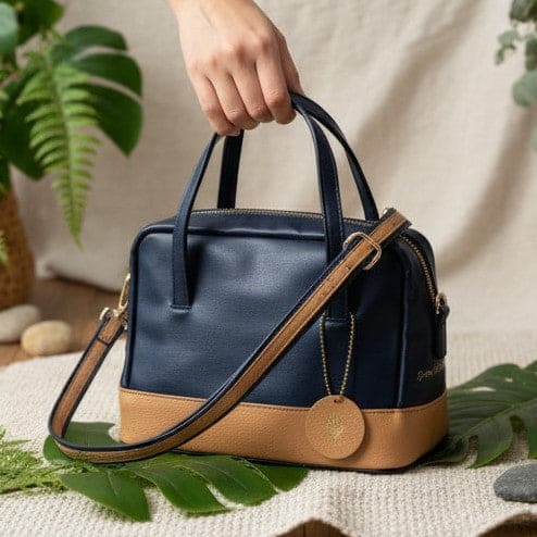 Navy blue and caramel plant leather satchel crossbody bag held by hand with strap visible, placed on a textured surface with green leaves, highlighting material and craftsmanship details