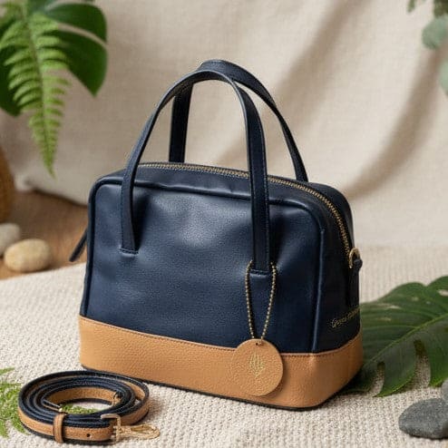 Navy blue and caramel plant leather satchel crossbody bag placed on a textured fabric surface with green leaves, showing front view with detachable strap and natural styling